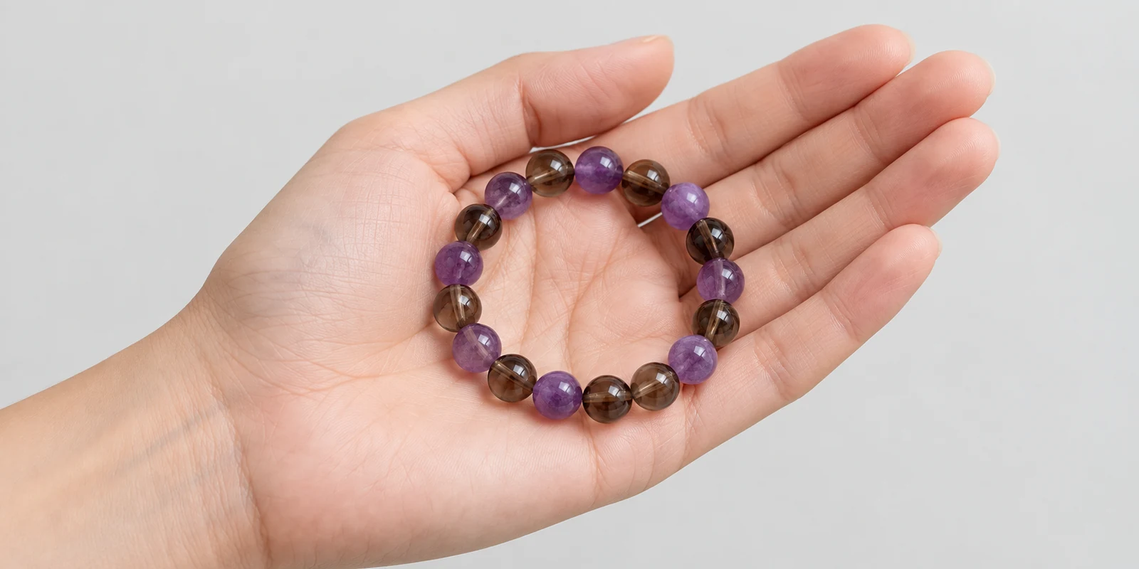Hand holding an amethyst and smoky quartz bracelet against a clean light background